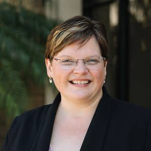Smiling woman with short hair and glasses wearing a black blazer, outdoors with greenery in the background.
