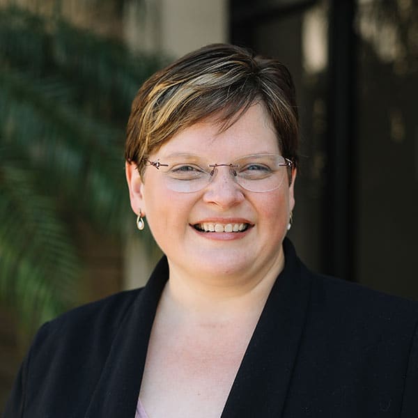 Smiling woman with short hair and glasses wearing a black blazer, outdoors with greenery in the background.