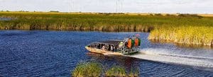 Motorboat with passengers wearing life jackets speeding through a grassy wetlands canal in a marshy landscape.