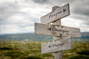 Wooden signpost with three boards reading 'Prayer', 'Changes', and 'Things' against a rural landscape.