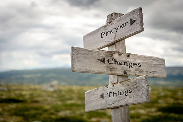 Wooden signpost with three boards reading 'Prayer', 'Changes', and 'Things' against a rural landscape.